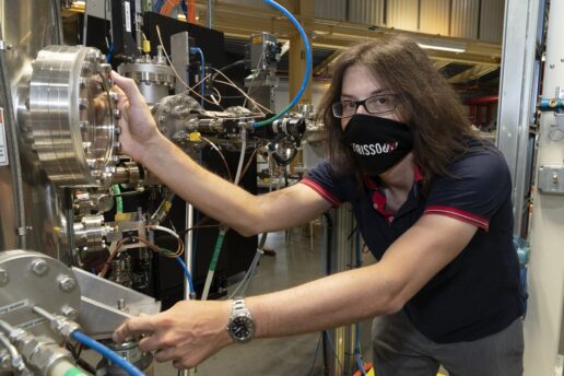 Photo of Jonathan Pelliciari in a lab, wearing a face mask and holding onto a large instrument with many metal knobs and wires.