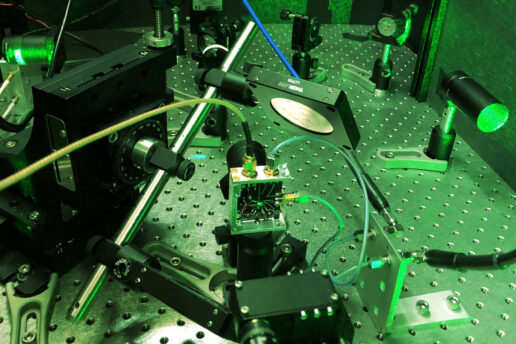 Photo of a tabletop experimental setup featuring lasers, mirrors, and other small, black or metal parts that are screwed into a large metal plate covered with screw openings. The entire scene is tinted green due to the green laser light being used.