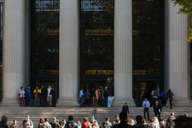 Photo of students crossing the street at 77 Mass. Ave., in front of MIT's main entrance, which features steps, large columns, and glass windows.