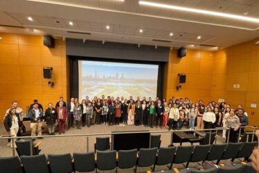 About 65 people pose standing in a semicircle on a large auditorium stage, with the backs of empty seats in the foreground and a video screen behind them.