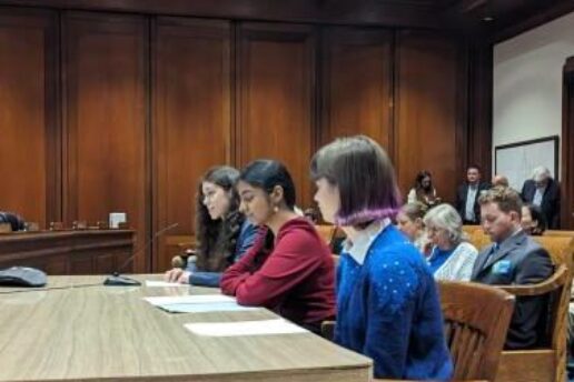 three students sit at desk with others sitting in gallery