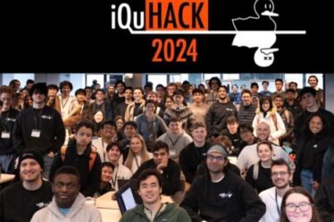 Perhaps 150 people pose for a photo in a large classroom. About a third are seated in the foreground and the rest stand. At top is a banner that reads iQuHACK 2024, with the image of two nearly identical ducks, one alive and one shown as dead with X's for eyes.