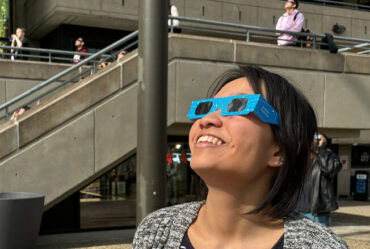 Joanna Chen wears special eclipse glasses and look up, with other people doing the same in background.