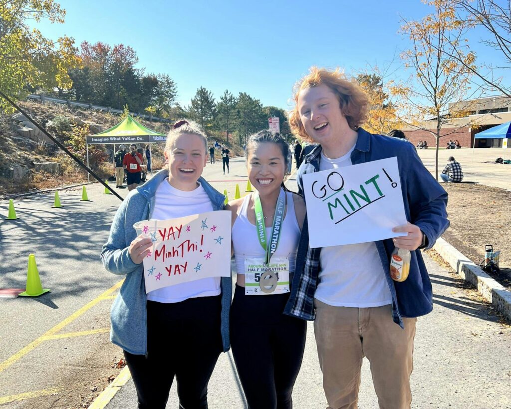 Two friends holding homemade signs flank Minh-Thi, congratulating her for finishing half marathon