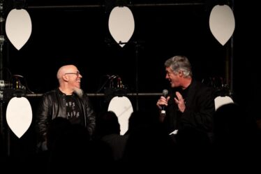 Jordan Rudess and Joseph Paradiso speak to each other on a black stage with white petals interspersed among them