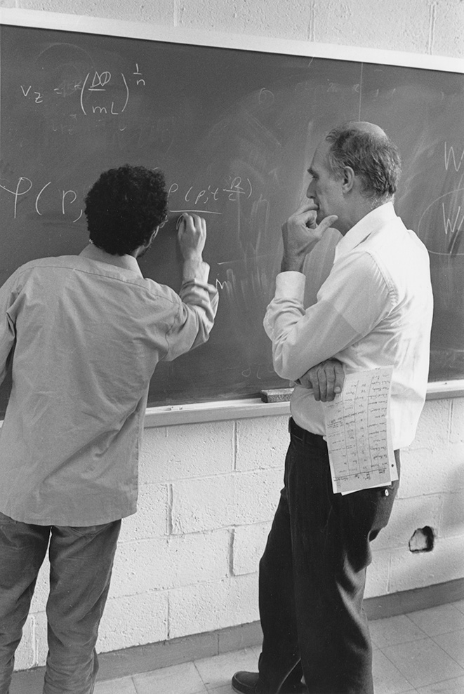 Physics looks on as student writes equation on blackboard